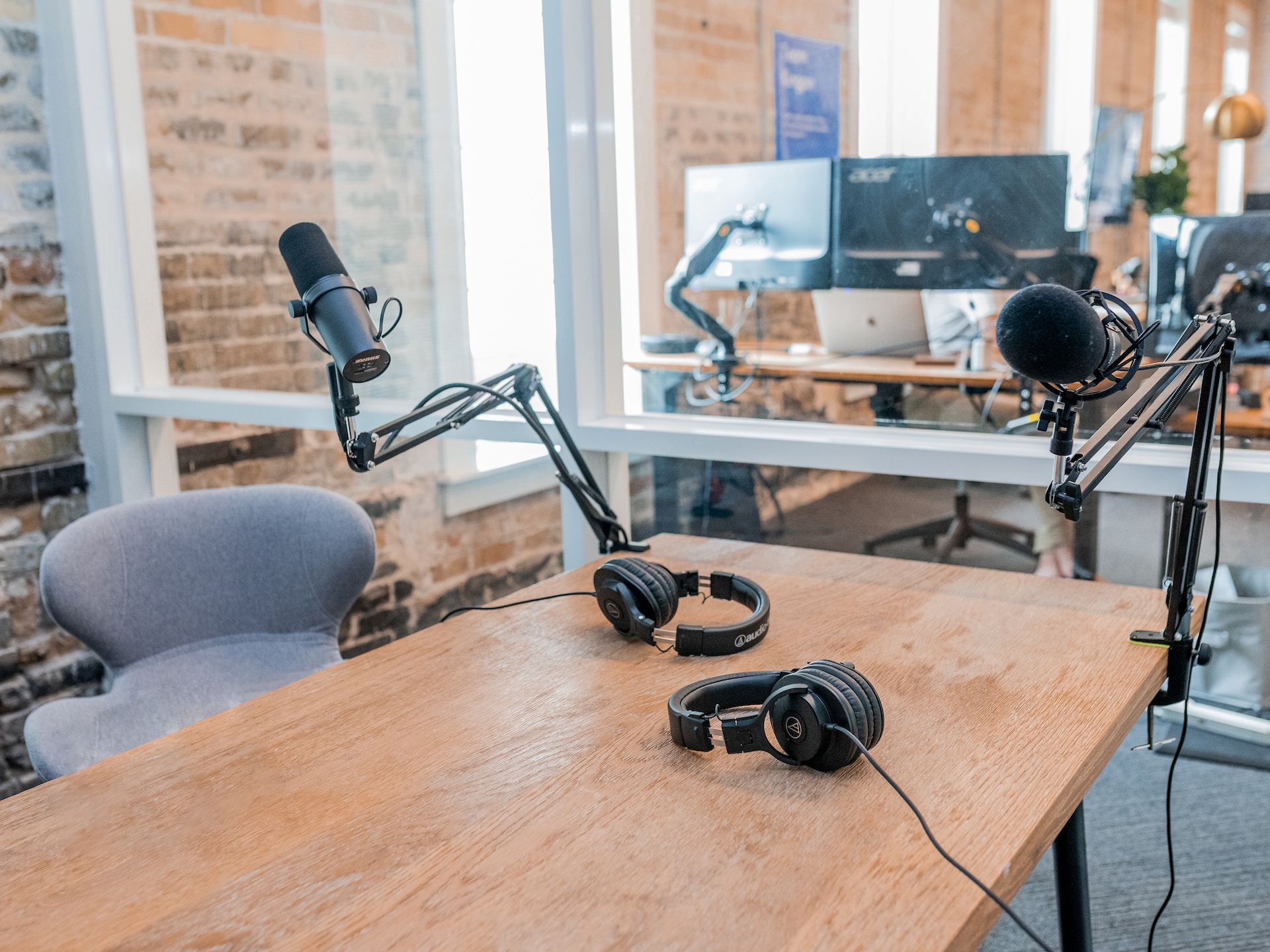 Photo of what appears to be an in-office podcast studio: a wooden table, with a microphone and set of headphones on either side.  An office environment is visible in the background, through a glass wall.  Photo by Austin Distel on Unsplash.