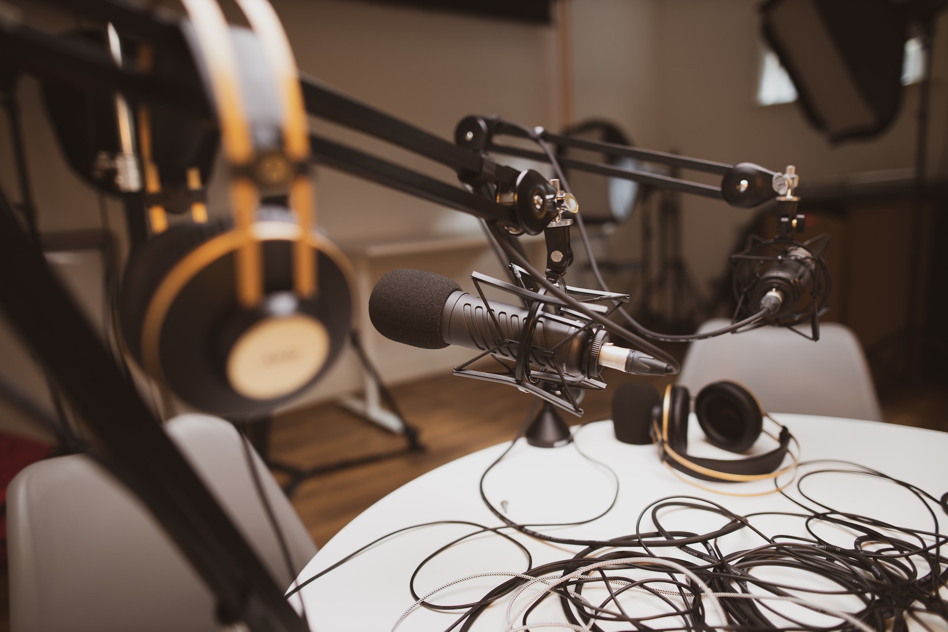 description: A podcast studio.  A microphone on a boom is in focus.  A set of headphones in the foreground, and the table in chairs in the background are out of focus.  Photo by Jonathan Farber on Unsplash.