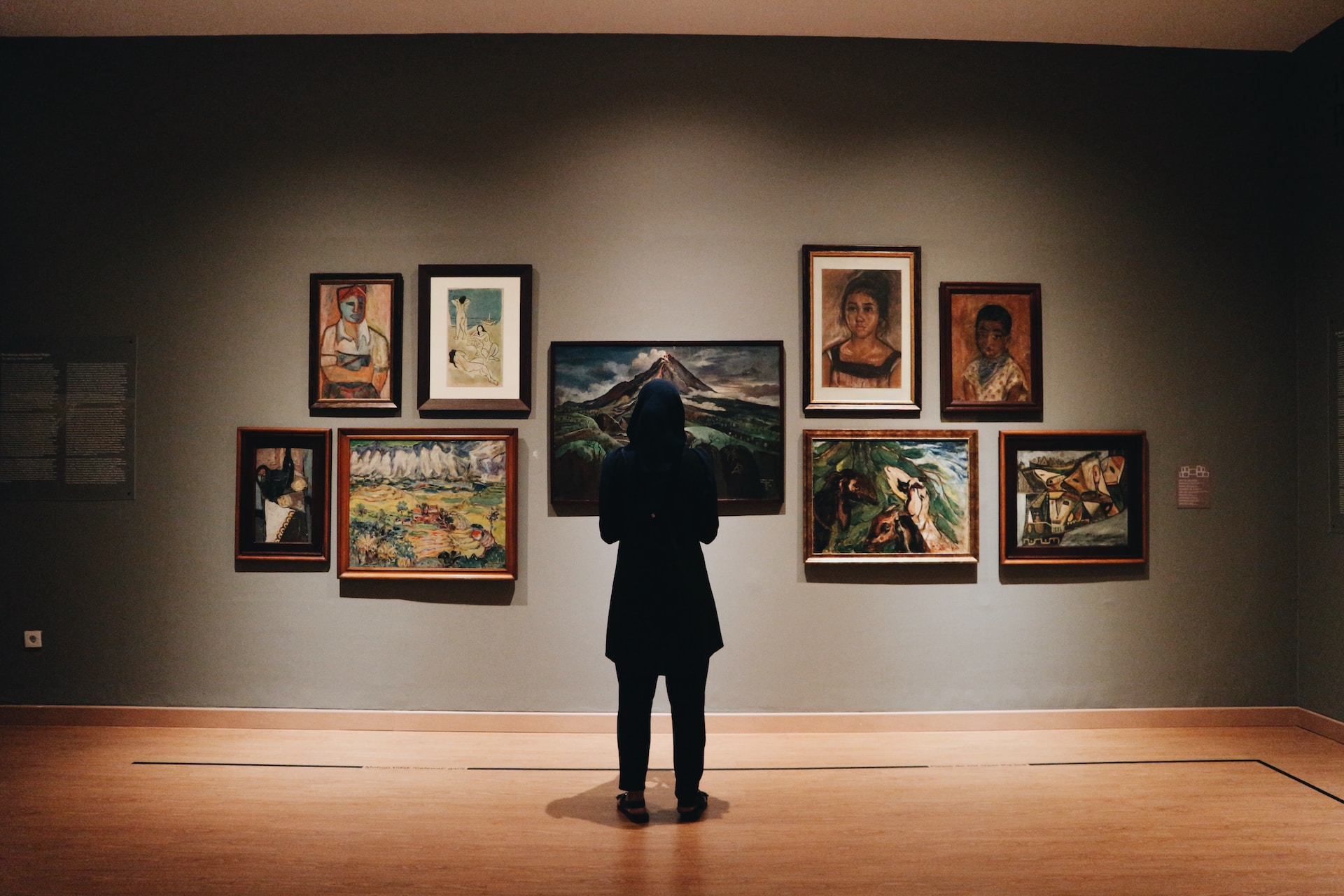 Description: A woman, in a black coat, has her back to the camera.  She is facing a wall of paintings in an art gallery.  Photo by Zalfa Imani on Unsplash.