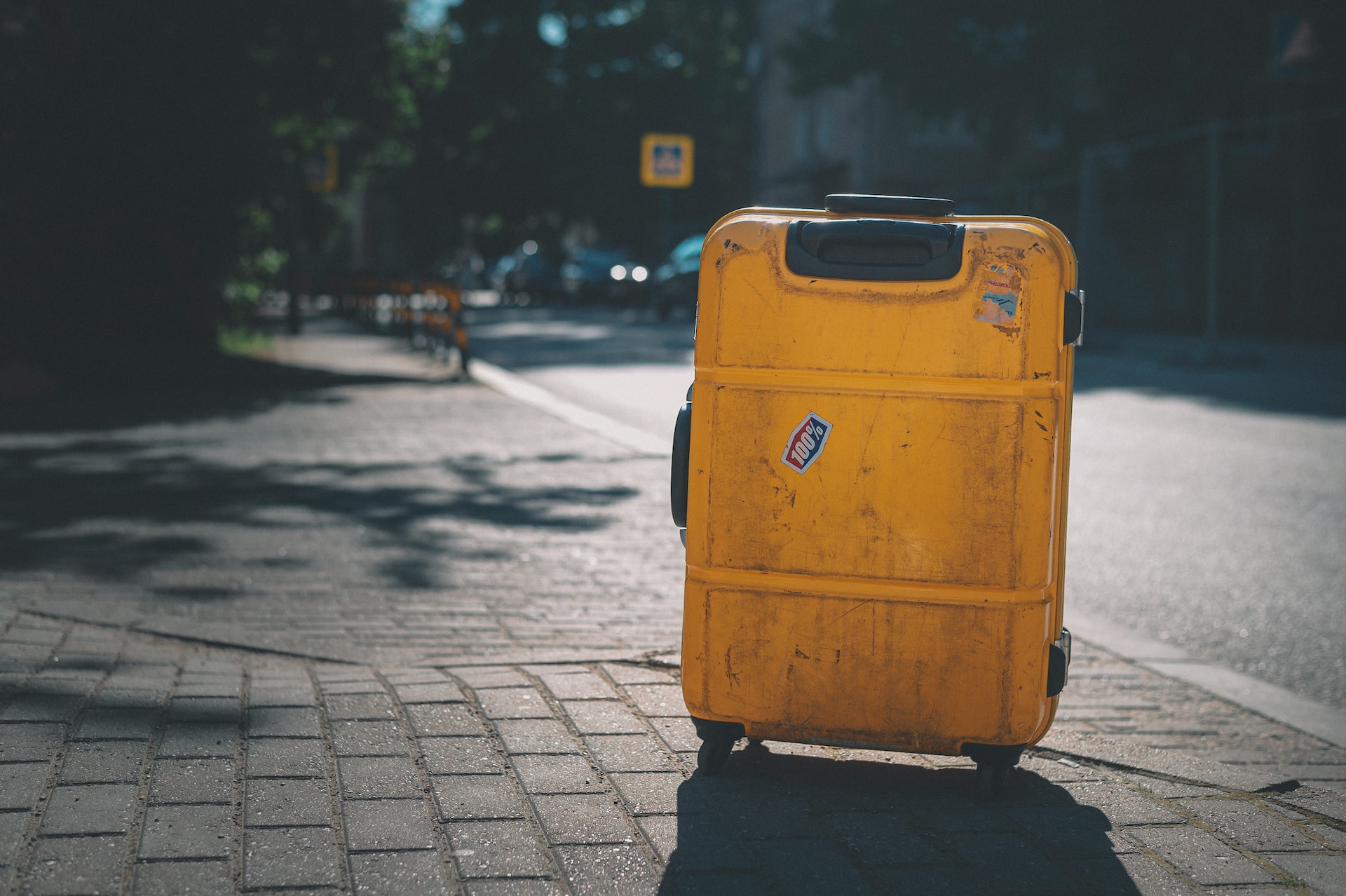 description: An image of a wheeled yellow suitcase, upright, on the sidewalk.  The backdrop of a city street is slightly out of focus.