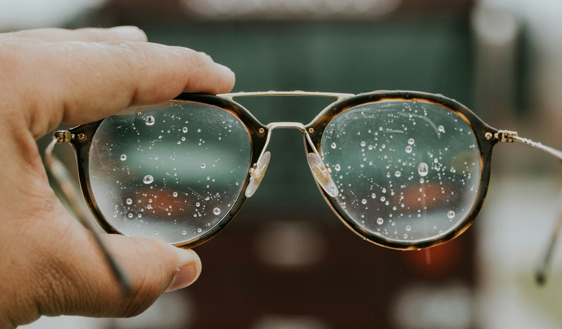 description: A hand holding up a pair of glasses.  The lenses are streaked with droplets, making it difficult to see through them. Photo by Hermes Rivera on Unsplash.